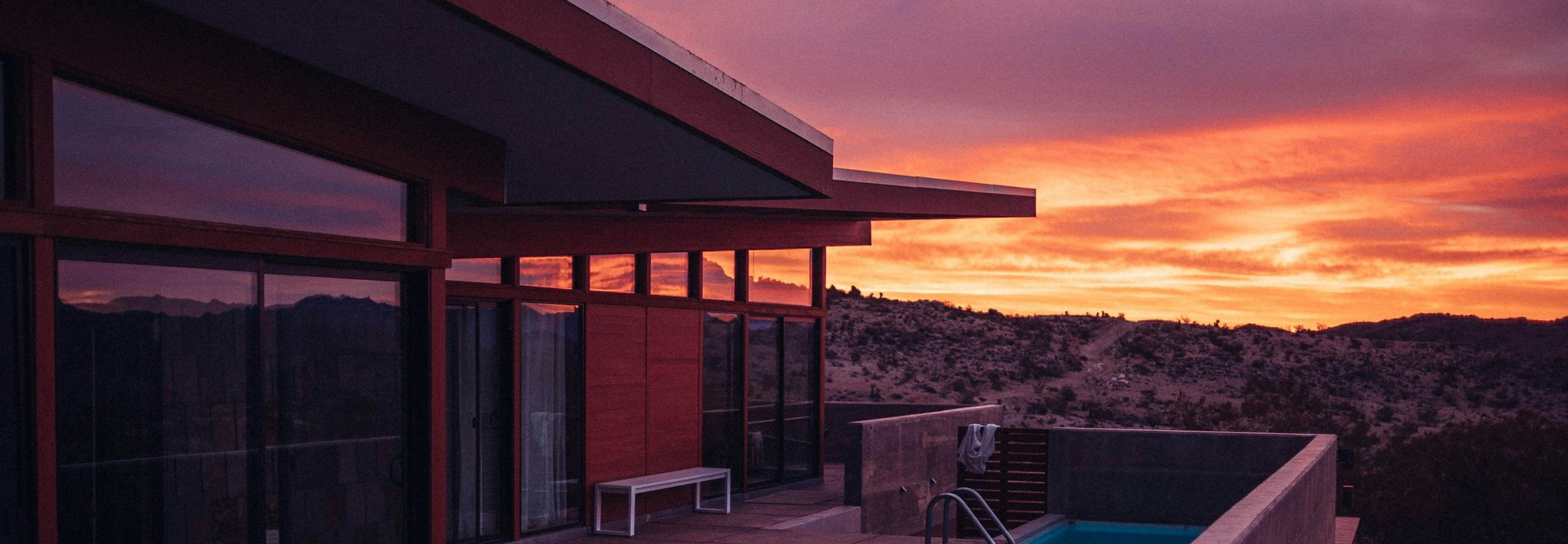 A midcentury modern house at sunset — glass walls reflecting the sky, a pool at its edge, hills beyond